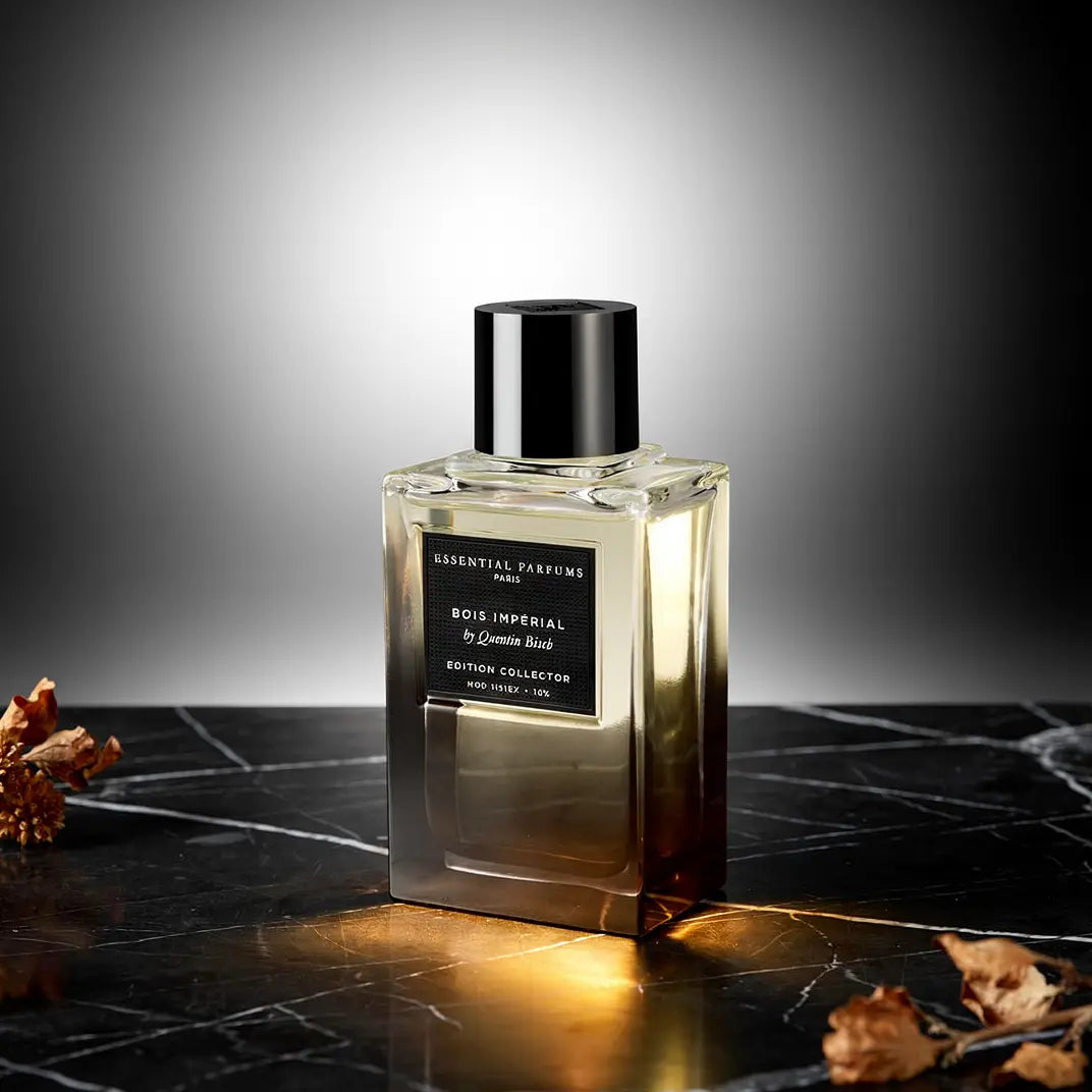 Flacon de parfum transparent avec bouchon noir, étiquette noir et doré, émanant une lueur chaude sur un plan noir marbré. Bois Impérial - Edition Collector 2025.